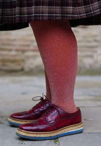 Jambes portant des collants rouges brillants à motifs et des chaussures brogues bordeaux polis avec des semelles épaisses en corde et caoutchouc, debout sur un pavé en pierre.