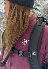 Veste en polaire bordeaux avec logo blanc, bretelles de sac à dos noires ajustables avec boucle, et un bonnet en tricot noir dans un décor forestier enneigé.