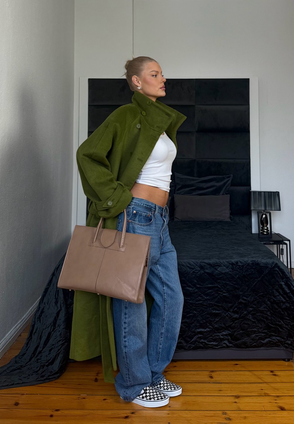 Femme en manteau vert, crop top blanc, jean bleu et baskets à carreaux tient un sac à main beige debout à côté d'un lit noir dans une chambre.