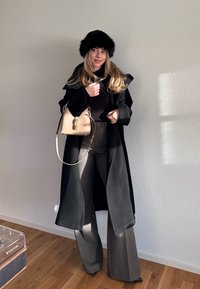 Kobieta w czarnym futrzanym kapeluszu, długim czarnym płaszczu i szarych szerokich spodniach, trzymająca beżową torebkę, stojąca na tle białej ściany.