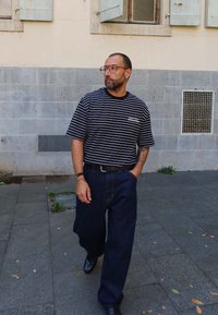 Mężczyzna z brodą i okularami, ubrany w pasiastą czarno-białą koszulę oraz szerokie ciemne dżinsy, idzie po brukowanej ulicy obok beżowej ściany.