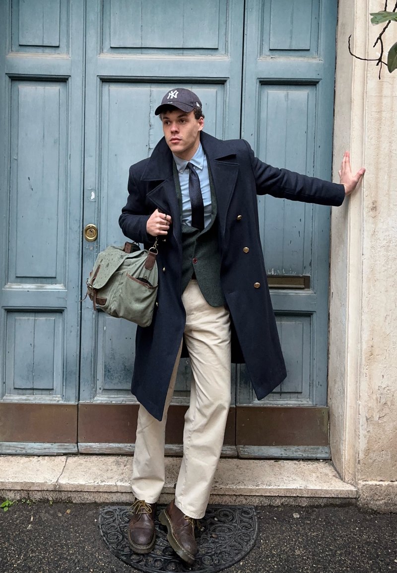 Jeune homme en manteau noir, chemise bleue, cravate, pantalon beige et casquette, debout à côté d'une porte bleue, tenant un sac vert et posant une main sur le mur.