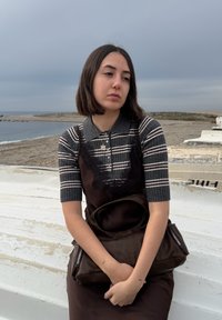 Νεαρή γυναίκα με ριγέ πουκάμισο και σκούρο φόρεμα κάθεται σε ένα λευκό σκάφος, κρατώντας μια καφέ τσάντα, με συννεφιασμένο ουρανό και παραλία στο φόντο.