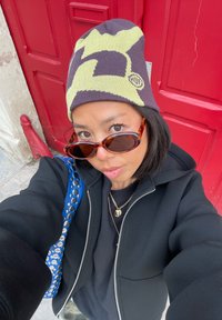 Bonnet tricoté à motif marron foncé et vert fluo, lunettes de soleil rectangulaires, veste noire zippée et sac à bandoulière bleu à motif devant une porte rouge.