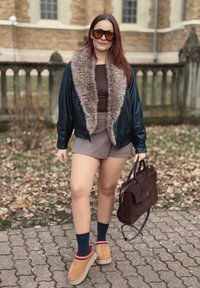 Černá kožená bomber bunda s límcem z umělé kožešiny, hnědý top, šedé šortky, vínová taška, světle hnědé boty s červenými akcenty a tmavě modré ponožky.