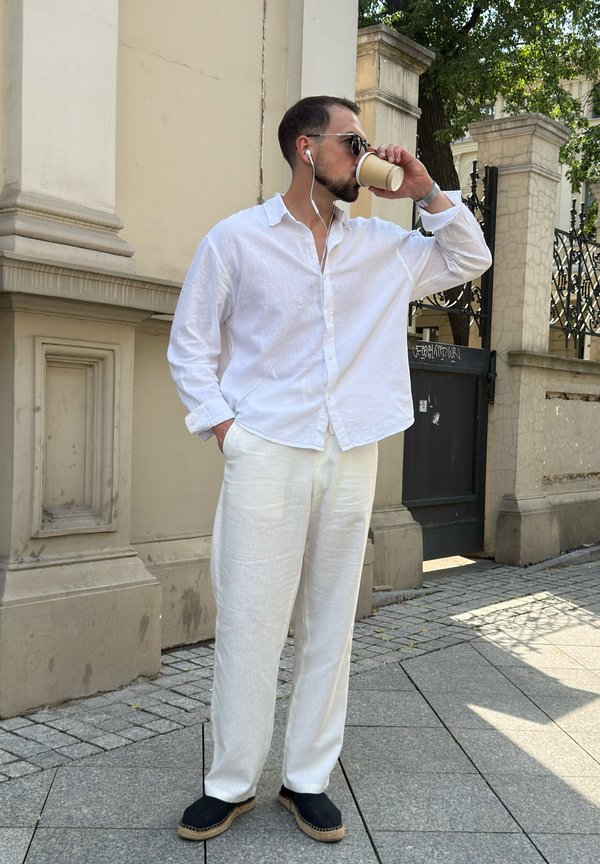 Homme portant une chemise et un pantalon blancs avec des chaussures noires, des lunettes de soleil et des écouteurs, buvant dans un gobelet en papier sur un trottoir en ville.
