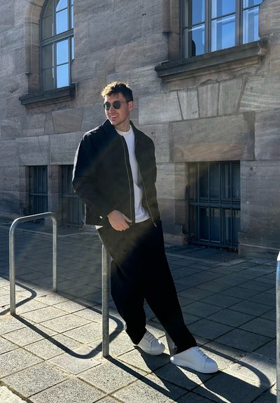 Chaqueta bomber negra sobre una camiseta blanca, combinada con pantalones negros holgados y zapatillas blancas. El hombre se apoya en una barandilla de metal.