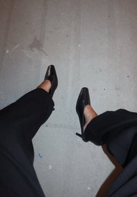 Schwarze High-Heels mit spitzer Spitze und offenen Seiten, kombiniert mit locker sitzenden schwarzen Hosen, auf einer einfarbigen grauen Oberfläche.