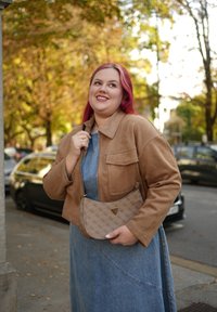 Brązowa zamszowa kurtka o skróconym kroju, zestawiona z sukienką z denimu chambray. Dodatki to beżowa torebka w wzory z logo.