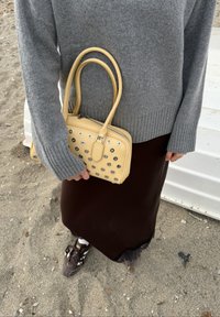 Άτομο που φοράει γκρι πουλόβερ και σκούρα φούστα σε αμμώδες έδαφος, κρατώντας μια μπεζ τσάντα χειρός με μεταλλικά τρυπήματα και φοράει καφέ αθλητικά παπούτσια.