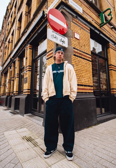 Conjunto informal con una sudadera beige con cremallera, camiseta gráfica azul, pantalones negros holgados y zapatillas de skate negras. Entorno urbano con paredes de ladrillo.