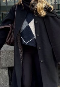 Long manteau noir avec de gros boutons, porté sur un pull géométrique texturé noir et gris. Sac à main tressé foncé ajoutant du détail. Pantalon noir.