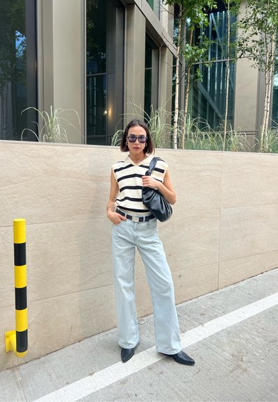 Top sin mangas a rayas en crema y negro, jeans de pierna ancha azul claro, bolso negro y zapatos negros de punta, en un entorno de un edificio moderno.
