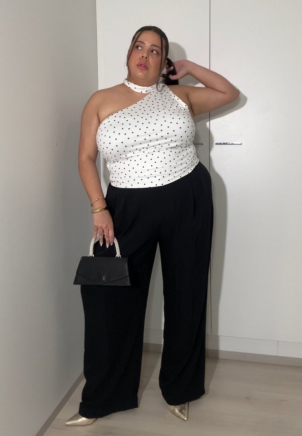 Woman in white polka-dot one-shoulder top and black wide-leg trousers holding black handbag with pearl handle, standing indoors.