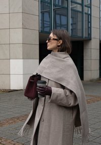 Manteau en laine beige avec un large col, porté sur un haut gris. Tenant une tasse à café noire et un sac en daim bordeaux. Gants en cuir marron.
