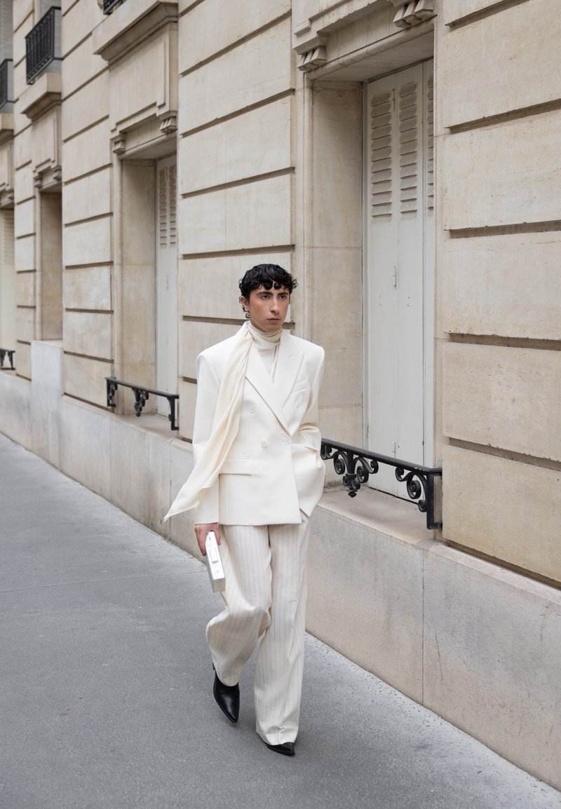 Person walking on city sidewalk wearing white double-breasted blazer, striped pants, scarf, holding a rectangular clutch, black pointed shoes.