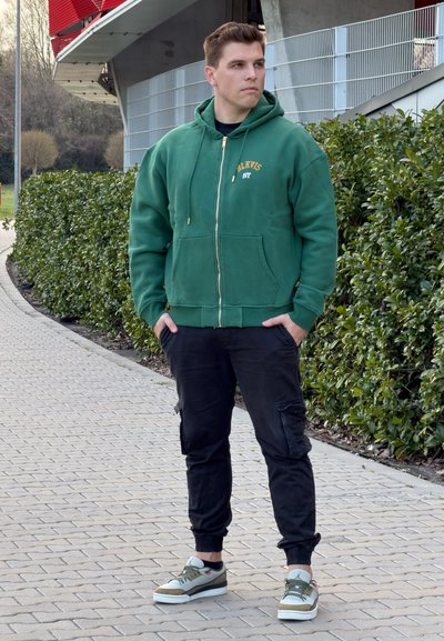 Joven con sudadera con capucha verde y pantalones cargo negros de pie en un camino pavimentado junto a un seto verde, mirando hacia un lado al aire libre.