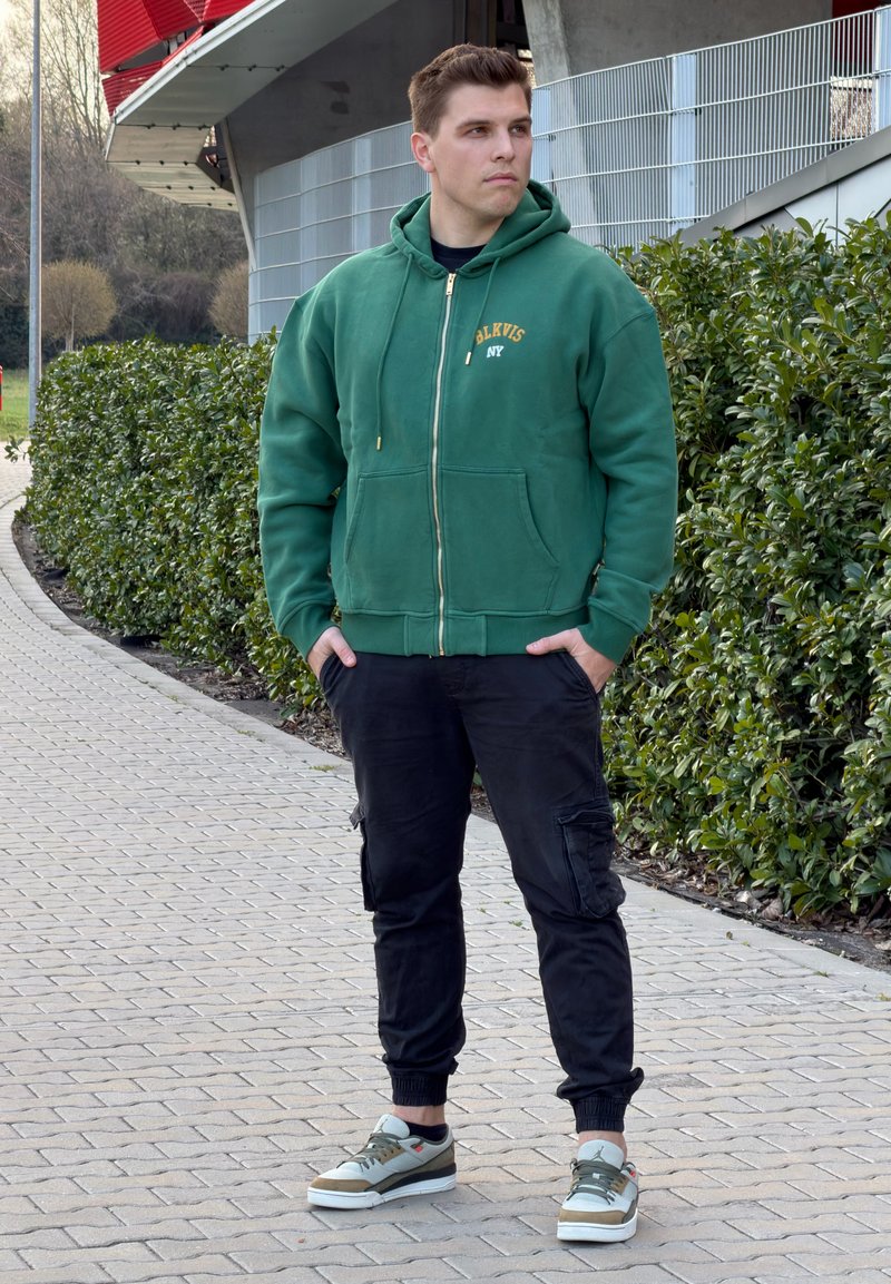 Jauns vīrietis zaļā kapučjakā un melnās cargo biksēs stāv uz bruģēta ceļa pie zaļas dzīvžoga, skatoties uz sāniem ārā.