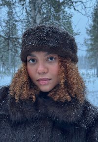 Femme aux cheveux bouclés portant un chapeau en fourrure foncée et un manteau, debout à l'extérieur avec des flocons de neige sur ses vêtements et des arbres en arrière-plan enneigés.