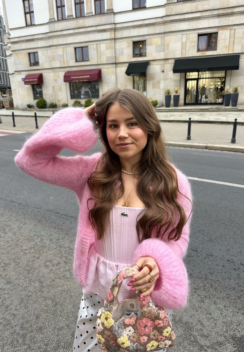 Jeune femme aux longs cheveux bruns portant un cardigan rose duveteux et un pantalon à pois, tenant un sac à main à motif floral dans une rue de la ville.