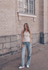 Top bianco senza maniche, jeans a gamba larga blu chiaro, sneakers bianche. Capelli lunghi e mossi di colore marrone. In piedi contro un muro di pietra testurizzato.