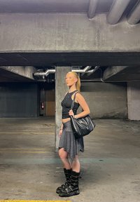 Top negru scurt cu pene, fustă evazată cu dungi, geantă neagră și bocanci masivi negri. Fundal din beton cu țevi la vedere.
