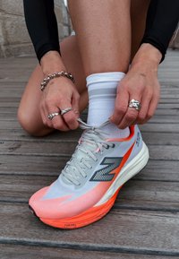 Svetlosivá bežecká topánka s jasne oranžovým detailom, sieťovaným zvrškom a odpruženou podrážkou. Šnúrky sa viažu, na pozadí je drevená podlaha.