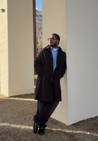 Abrigo marrón sobre una camisa azul claro, pantalones oscuros y zapatos negros, con tela texturizada y detalles de diseño a medida. De pie contra una pared.