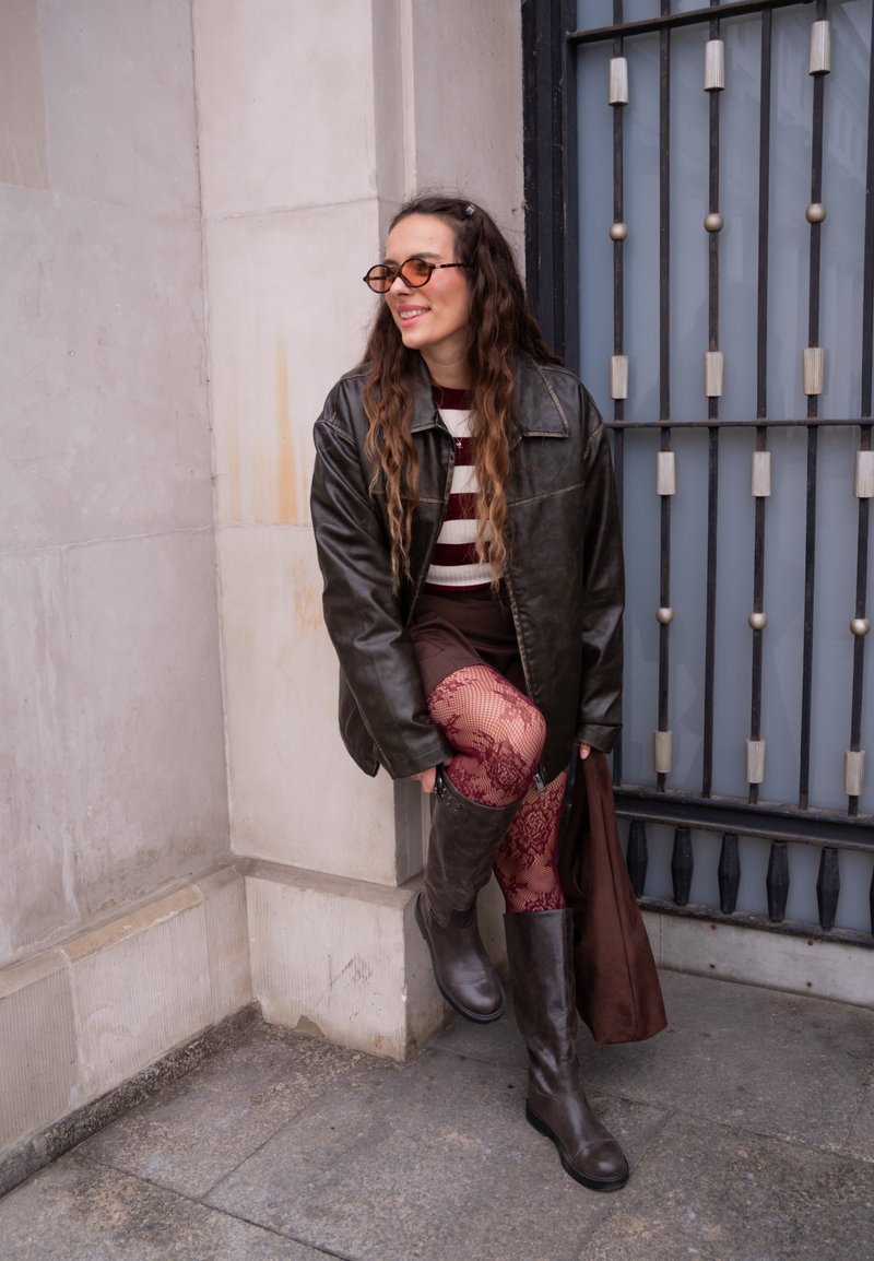 Brown leather oversized jacket, striped sweater, brown skirt, red lace fishnet tights, and knee-high brown boots. Brown tote bag beside.