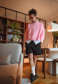 Homme aux cheveux bouclés et lunettes portant un pull rose, une chemise blanche, un short noir, et des sandales avec des chaussettes dans un salon moderne.