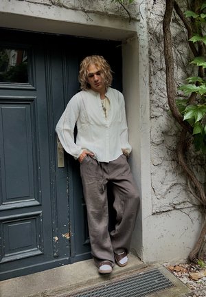 White cotton long-sleeve shirt with a relaxed fit, pleats, and a buttoned placket. Paired with loose grey linen trousers and brown sandals.