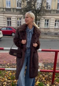 Długi brązowy płaszcz z futra sztucznego, dżinsowa koszula z zapięciem na muszkę, szerokie dżinsy i mała brązowa torebka, na tle ulicy.