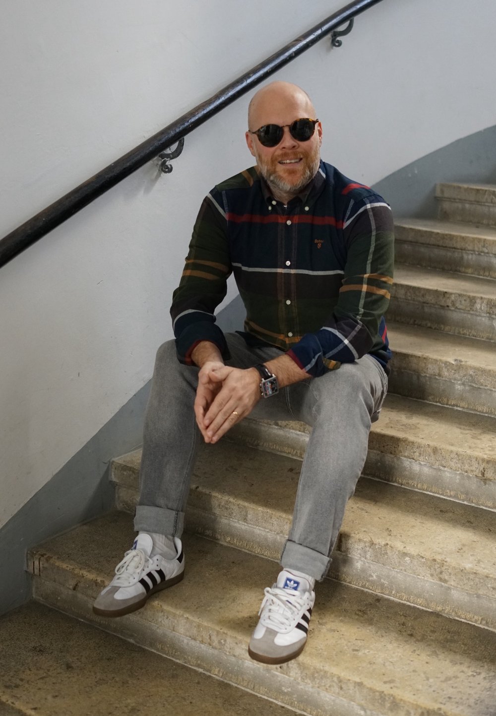 Camisa xadrez com riscas verdes, azuis, vermelhas e brancas; jeans cinza claro; sapatilhas brancas com riscas escuras; sentado em escadas de concreto.