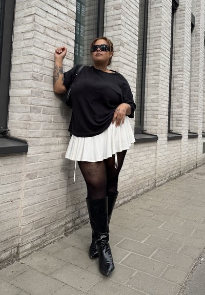 Mujer con camiseta negra de gran tamaño, falda blanca, medias negras y botas hasta la rodilla se apoya en una pared blanca de ladrillo en una acera pavimentada.