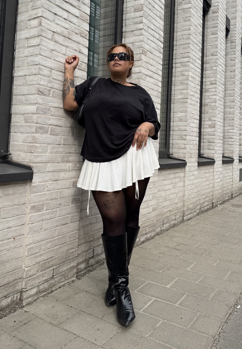 Donna che indossa una t-shirt nera oversize, gonna bianca, collant neri e stivali al ginocchio appoggiata a un muro di mattoni bianco su un marciapiede lastricato.