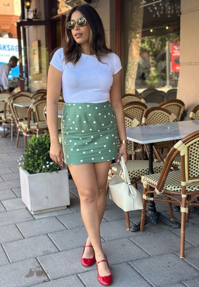 Camiseta blanca de canalé, falda verde con lunares blancos, zapatos tipo Mary Jane rojos y un bolso blanco; café al aire libre con sillas de fondo.