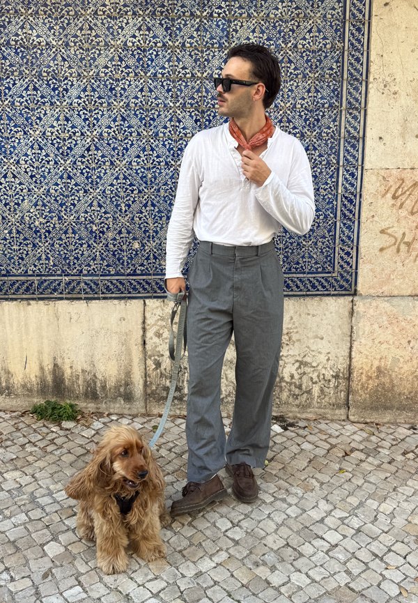 Homme en lunettes de soleil, chemise blanche, pantalon gris tenant la laisse d'un chien brun assis dans une rue pavée près d'un mur carrelé bleu à motifs.