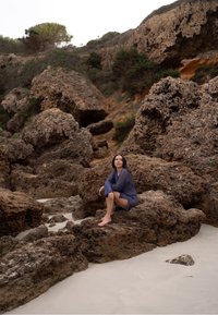 O persoană stă pe stânci ruginii texturate, în apropierea unei plaje cu nisip, purtând o pelerină tricotată de culoare bleumarin, cu mâneci lungi și o croială lejeră.