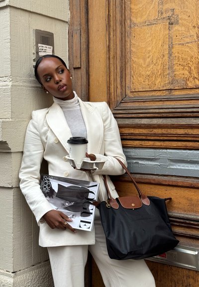 Blazer blanco y pantalones a juego, suéter de cuello alto gris, sosteniendo una taza de café, una revista y un bolso negro con detalles marrones, frente a una puerta de madera.