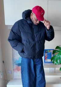 Čierna nadýchaná bunda so zapínaním na zips a kapucňou, skombinovaná so širokými modrými džínsami a červenou čiapkou. Vnútorné prostredie s viditeľnými rastlinami.