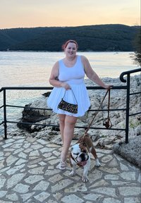 Weißes ärmelloses Kleid mit schwingendem Rock, kombiniert mit weißen Sandalen. Hält eine Handtasche mit schwarz-leopardenmuster und steht neben einem braunen Bulldoggen.
