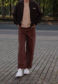 Braune zip-up Jacke über einem beigen Shirt, kombiniert mit weiten braunen Hosen und weißen Sneakers. Der Hintergrund zeigt einen gepflasterten Weg und Bäume.