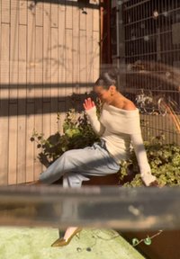 Off-white, geripptes Top ohne Träger, hellblaue weite Jeans, grüne Stilettos, sitzend zwischen Blumentöpfen, mit einer Holzwand im Hintergrund.