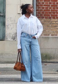 Biała koszula z kołnierzykiem, jasnoniebieskie jeansy o szerokich nogawkach oraz brązowa zamszowa torebka. W tle znajduje się teksturowana ściana z detalami z cegły.