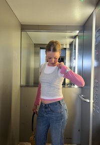 Femme en manche longue rose et débardeur gris ajustant sa manche à l'intérieur d'un petit ascenseur aux parois métalliques et muni d'un miroir.