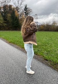 Bruine jas met schapenvachtvoering en een hoekig ontwerp, gecombineerd met lichtblauwe spijkerbroek en crèmekleurige sneakers. Gras en bomen op de achtergrond.