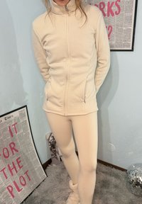 Jopica iz flisa v krem barvi z zadrgo, visokim ovratnikom, stranskimi žepi in oprijeto zasnovo, združena z ujemajočimi legicami. Gladek tekstura.