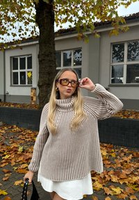 Bruin en crème gestreepte gebreide trui met een hoge kraag, gecombineerd met een witte jurk. Lange golvende haren, grote bruine zonnebril en een zwarte tas.