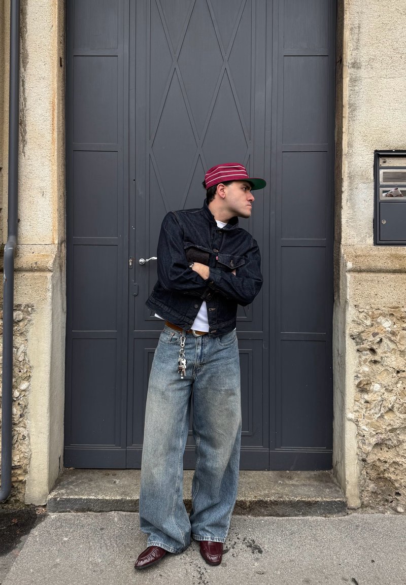 Homme portant une casquette rouge, une veste foncée et un jean large, debout les bras croisés, appuyé contre une porte grise dans un environnement urbain en pierre.
