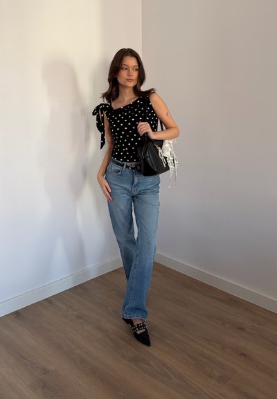 Joven mujer con blusa negra de lunares, jeans azules y zapatos negros puntiagudos, de pie sobre suelo de madera sosteniendo un bolso negro con detalle de encaje blanco.